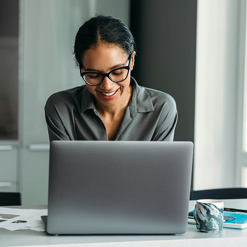 A woman wearing glasses and a greyish green shirt sits at a desk, her hair tied back, looking down at her computer with a gentle smile. 