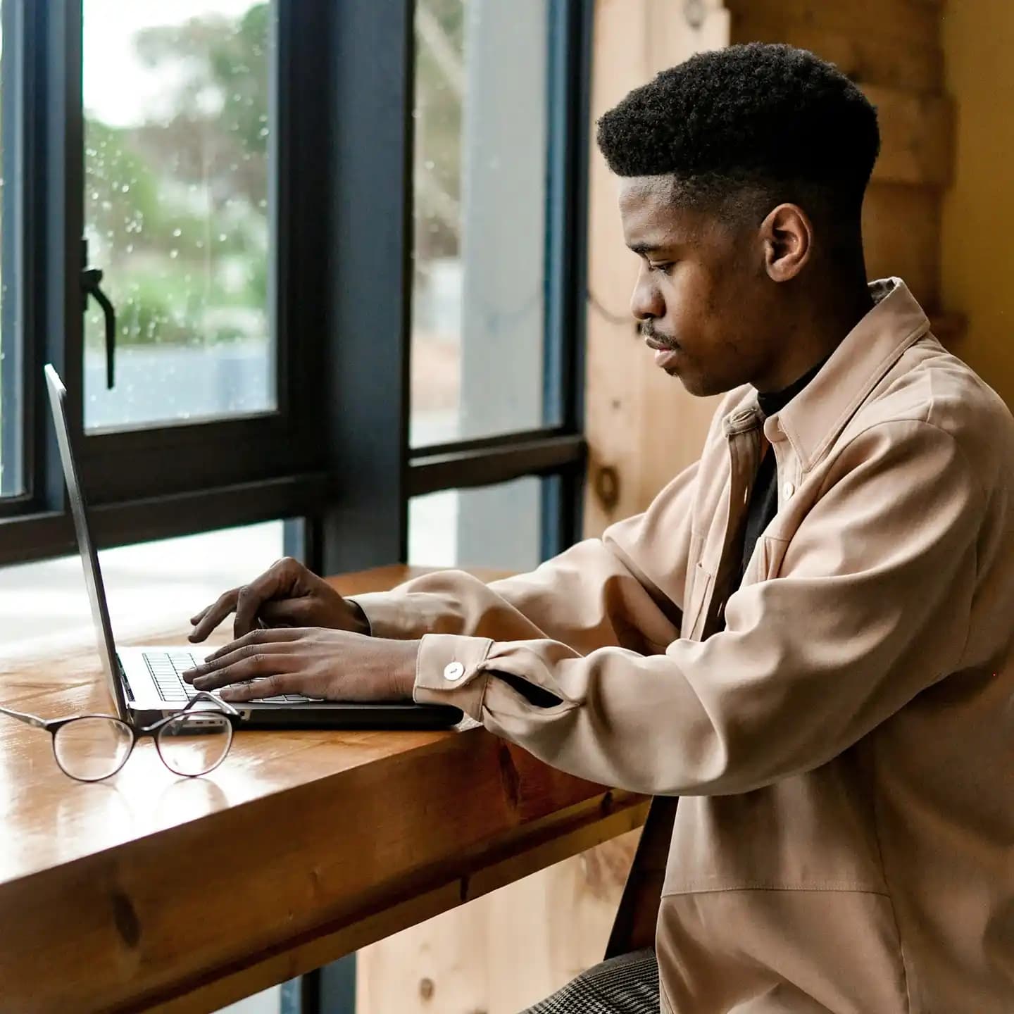 A young man in a light brown jacket focuses on his laptop while sitting at a table in front of a rainy window.
