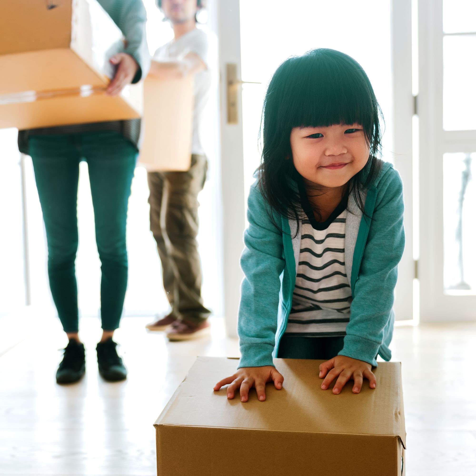A young girl with bangs wearing a teal jacket places her hands on a cardboard box and pushes it forward, while in the background, her parents’ legs are visible as they carry boxes, suggesting the family is moving in.