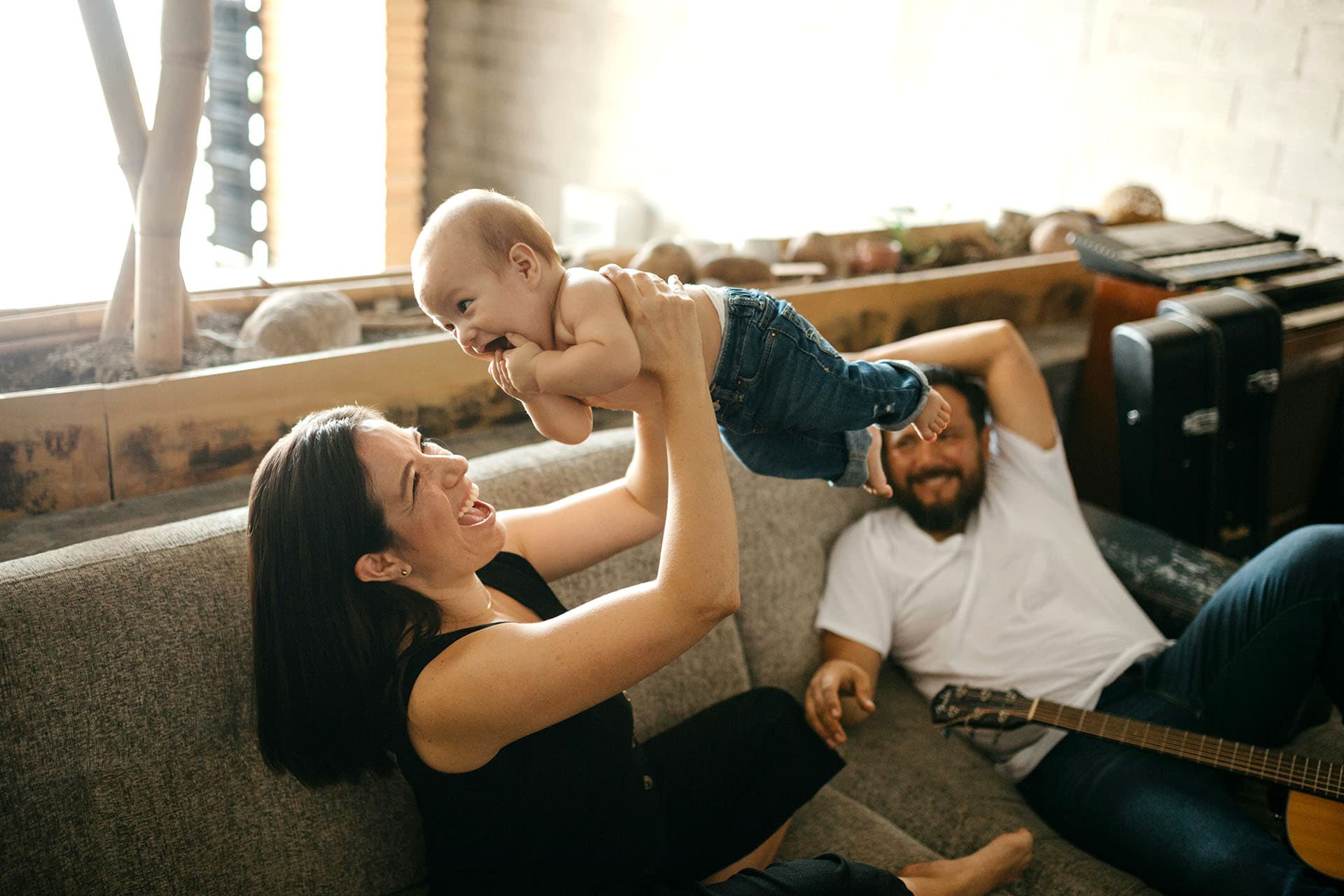 A family on the couch, with a woman smiling as she lifts a baby into the air, while the father lies back with a guitar, watching them happily.