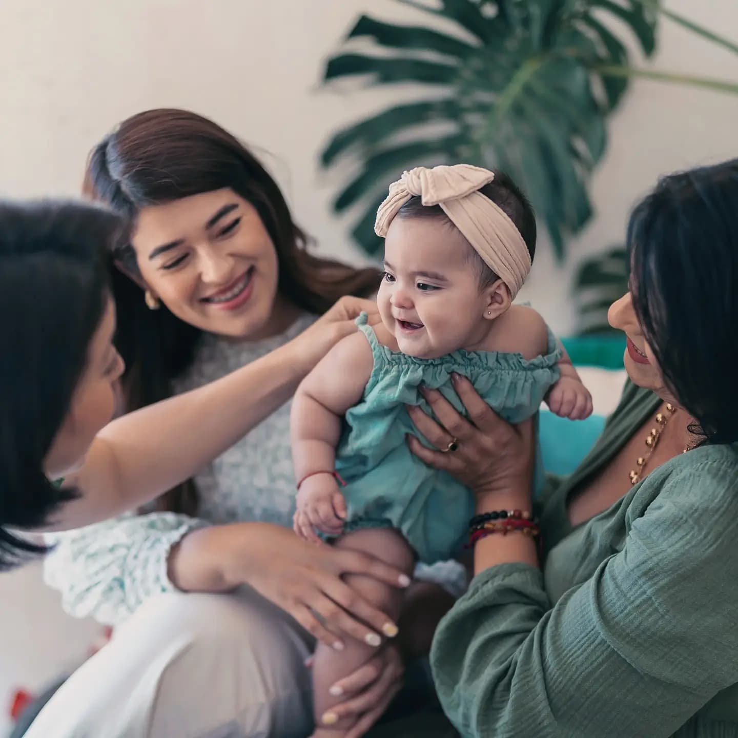 A baby dressed in teal clothes, surrounded by three smiling women who are all holding and looking lovingly at the baby.