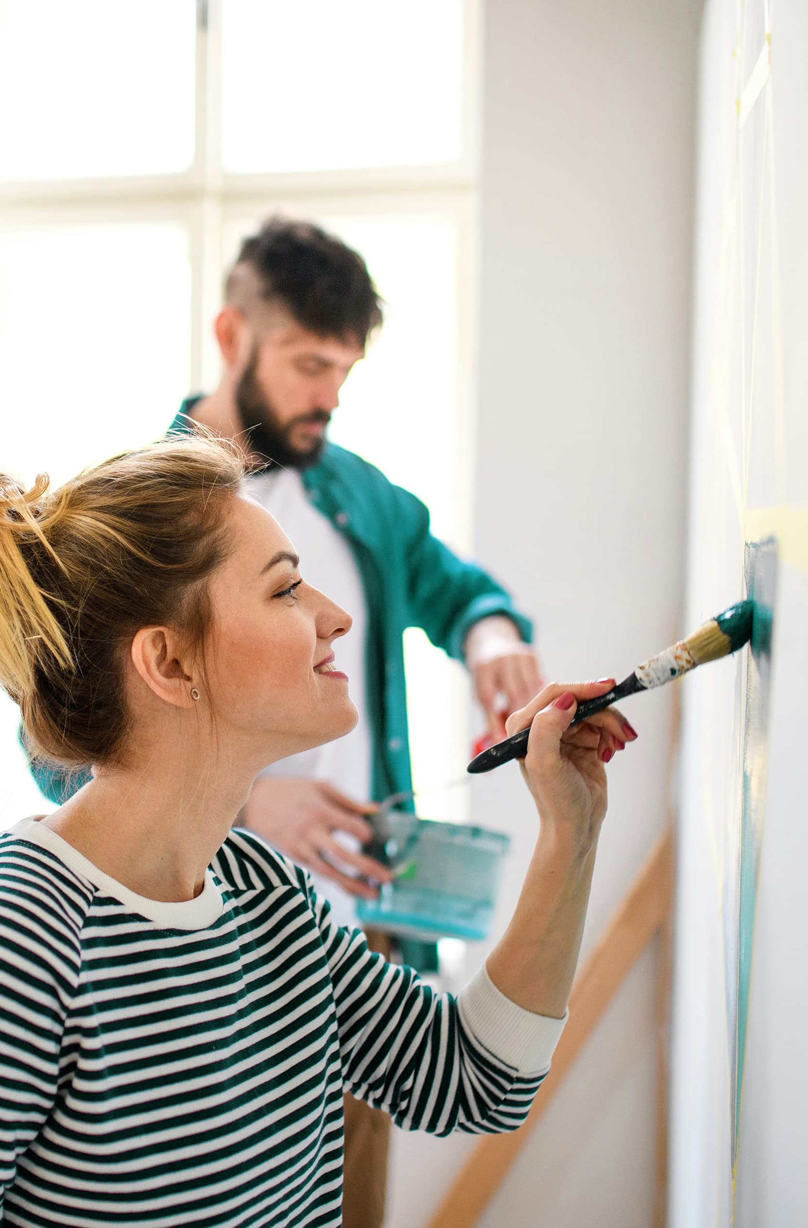 A close-up of a blonde woman painting a white wall with teal paint, wearing a striped shirt, while in the blurred background, a man in a teal shirt layered over a white shirt is visible.
