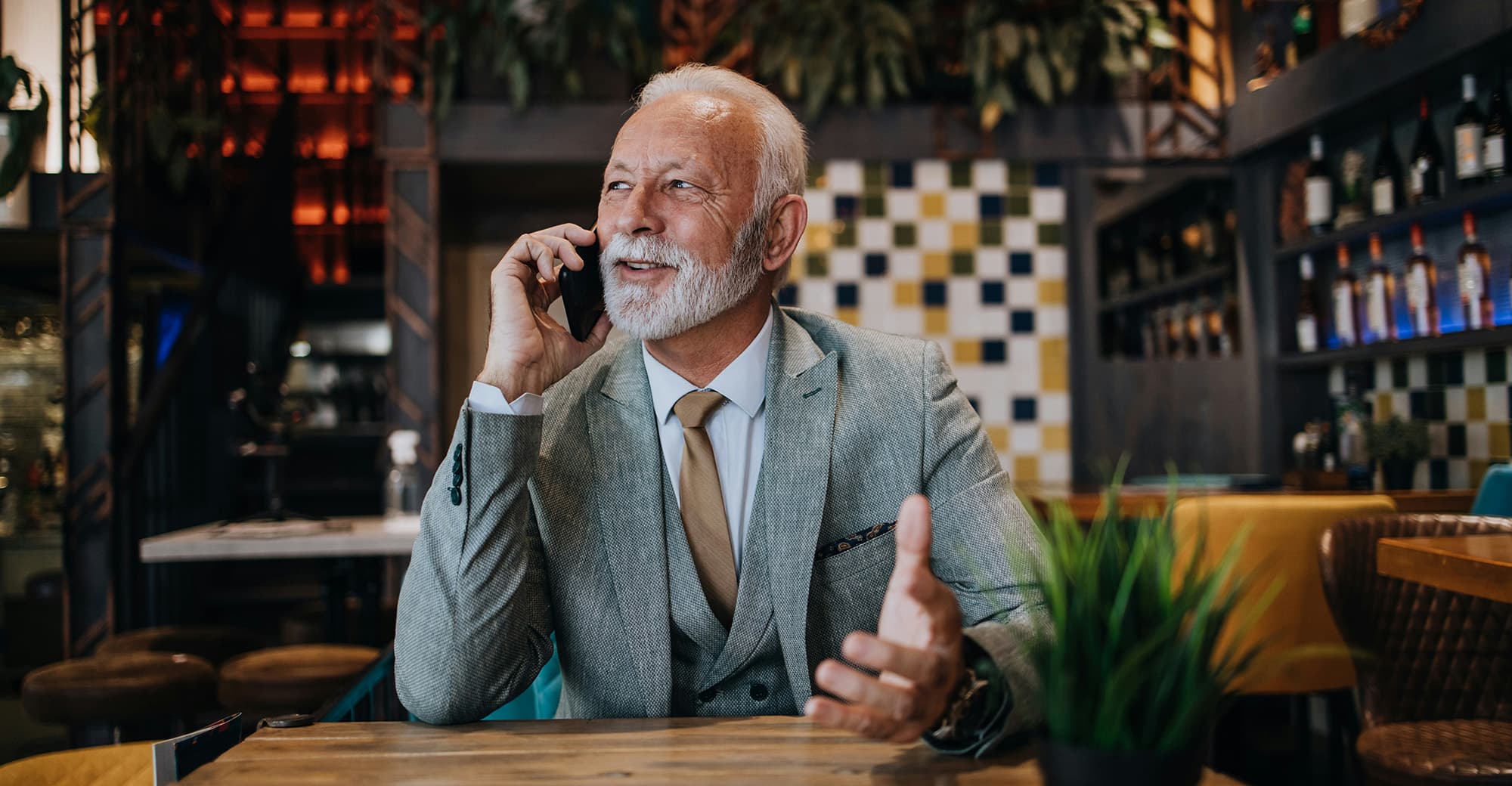 An elderly man with white hair, dressed in a grey suit and golden necktie, sits in a colourful restaurant, talking on the phone while looking to the side smiling.