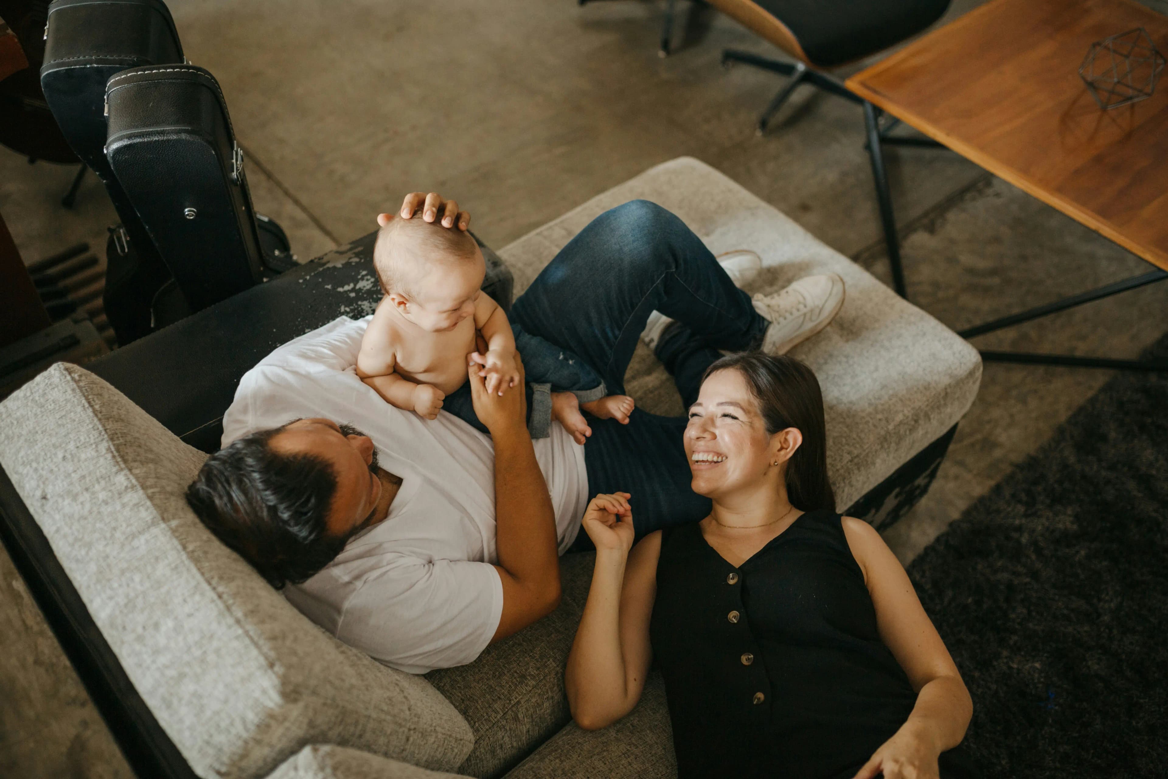 A warm family scene at home with the dad lying on a reclining chair holding a baby, while the mother laughs and rests her head on the chair.