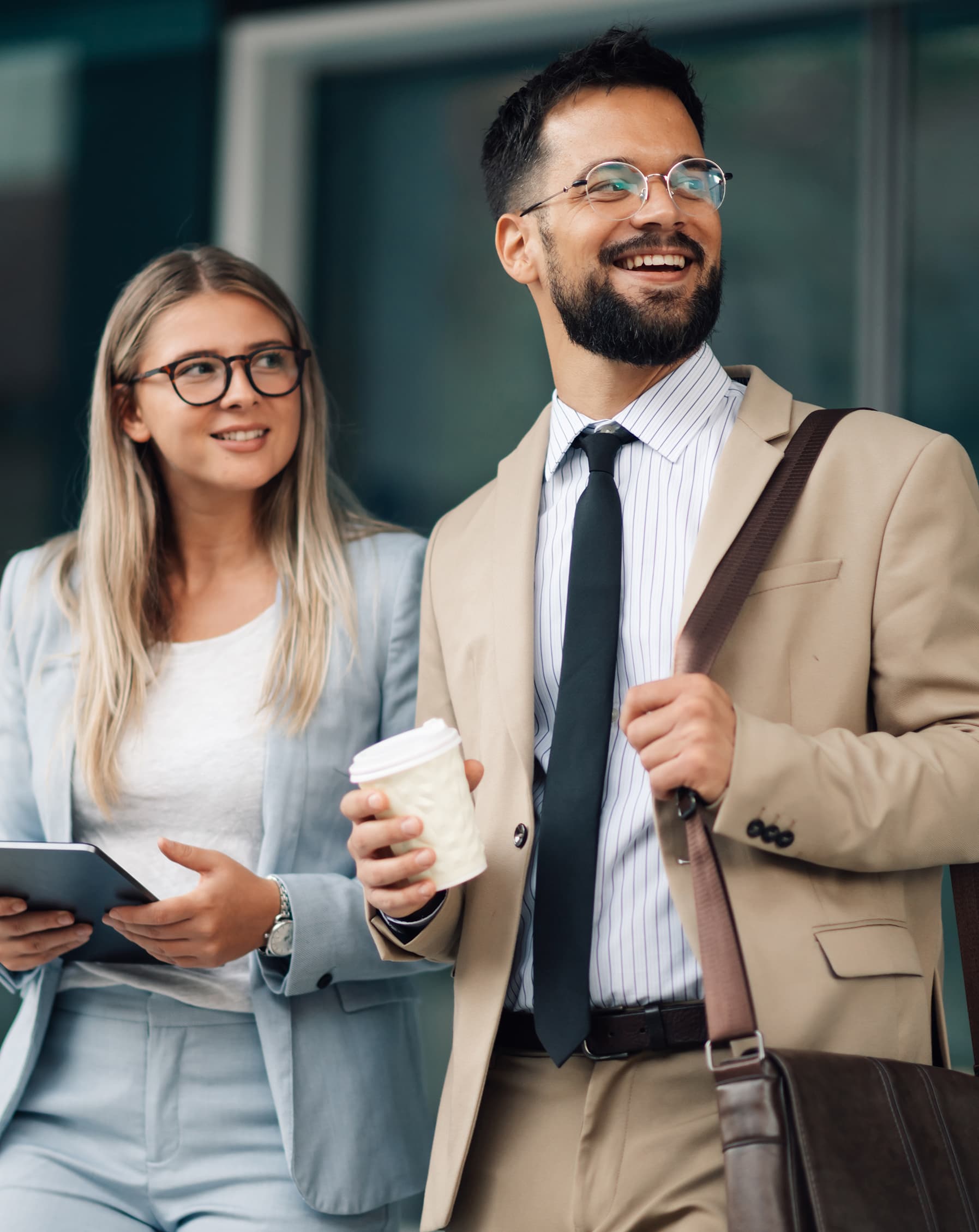 Two young business professionals are leaving the office after a productive day, holding coffee and a tablet and smiling at something in the distance