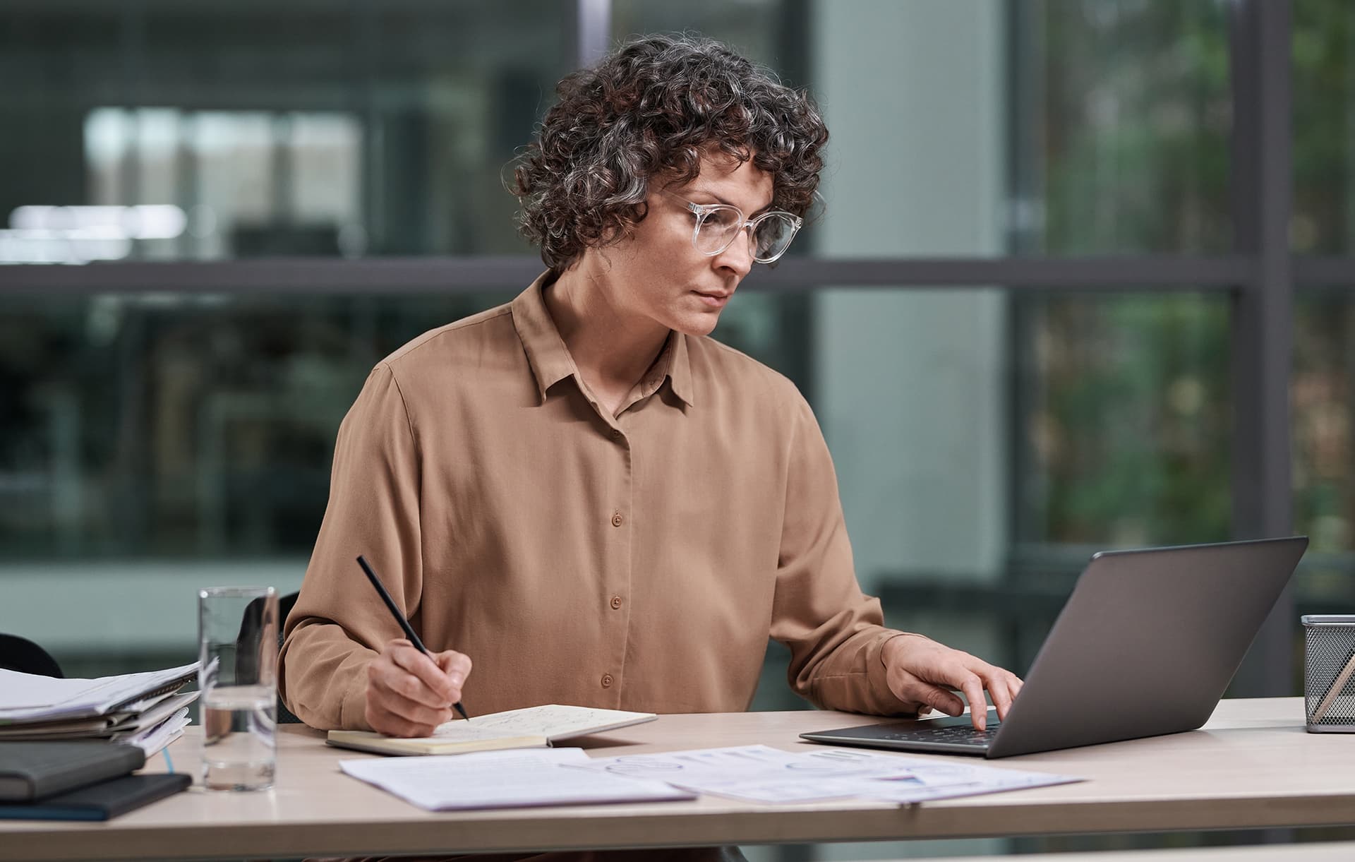 Experienced female broker in beige blouse working over business project while sitting by workplace in front of laptop and analyzing data