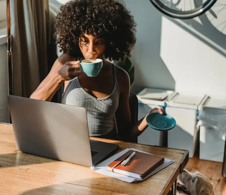 A woman working at a desk, drinking coffee while using a laptop, with a notebook and pen on the table, and a dog partially visible in the background.