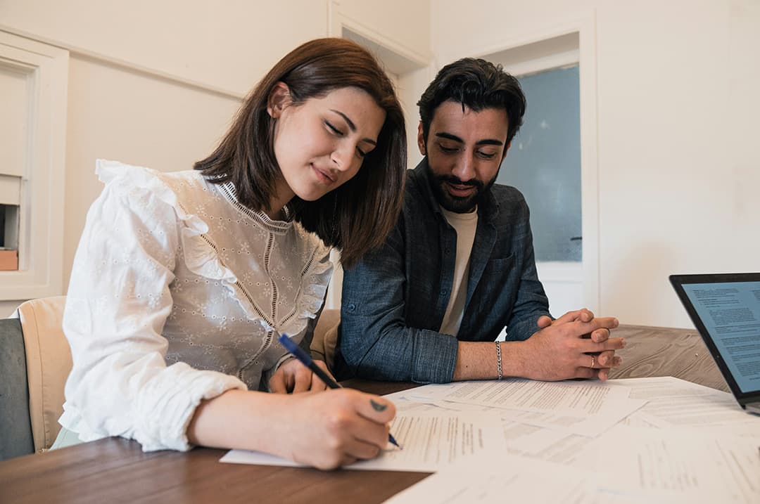 A woman and man sitting side by side at a table, both smiling as the woman signs a document and the man looks at the papers.