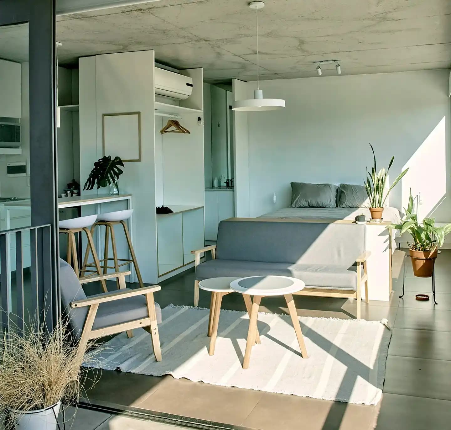 A well-lit living room that resembles a studio apartment, featuring a bed, compact kitchen, dining area, and several green plants.